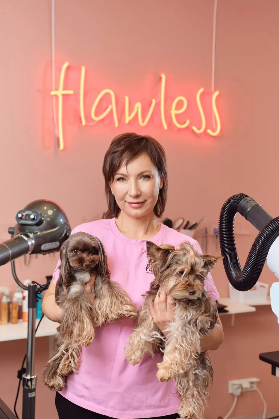 A groomer holding two Yorkshire terriers under the salon's neon sign